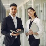 Confident well dressed multiracial coworkers standing in modern office corridor while holding laptop and notebook in hands and looking at camera